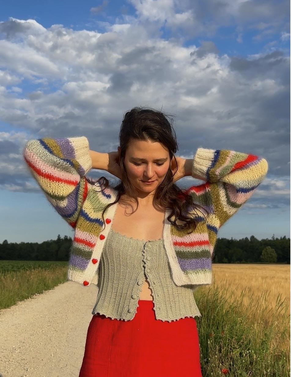 Lotti smiling outdoors in a wheat field, wearing a colourful striped mohair cardigan with heart-shaped buttons