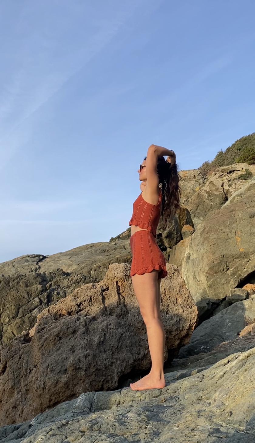 Rust-coloured hand-knitted crop top and shorts, worn on coastal rocks at sunset